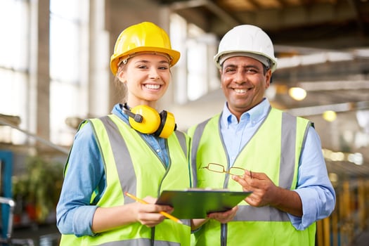 Two workers wearing hard hats and safety vests looking at a clipboard